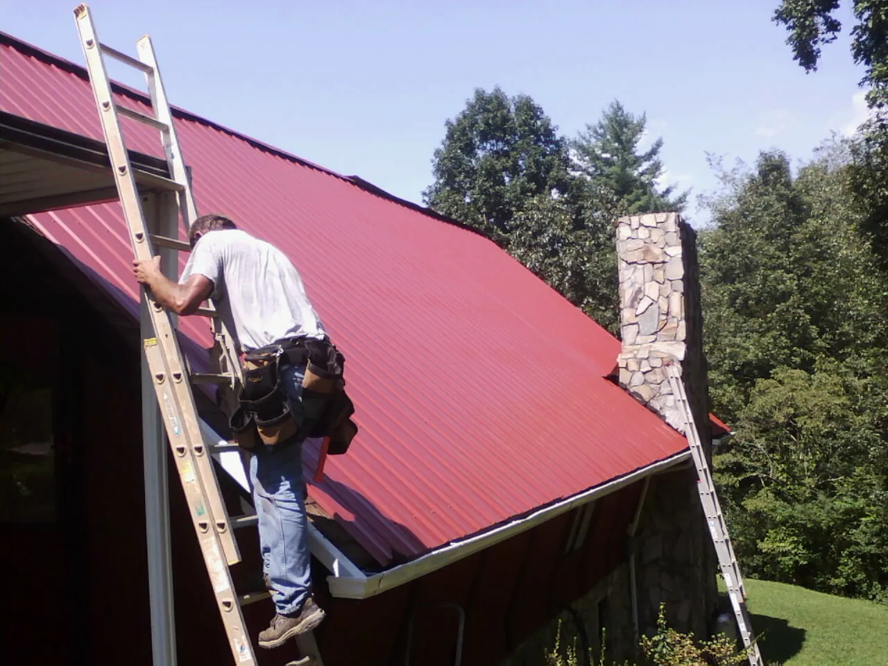 Roofer on a ladder beside a red metal roof and stone chimney — Soffit & Fascia Repair by Roofing Brothers in Bristol